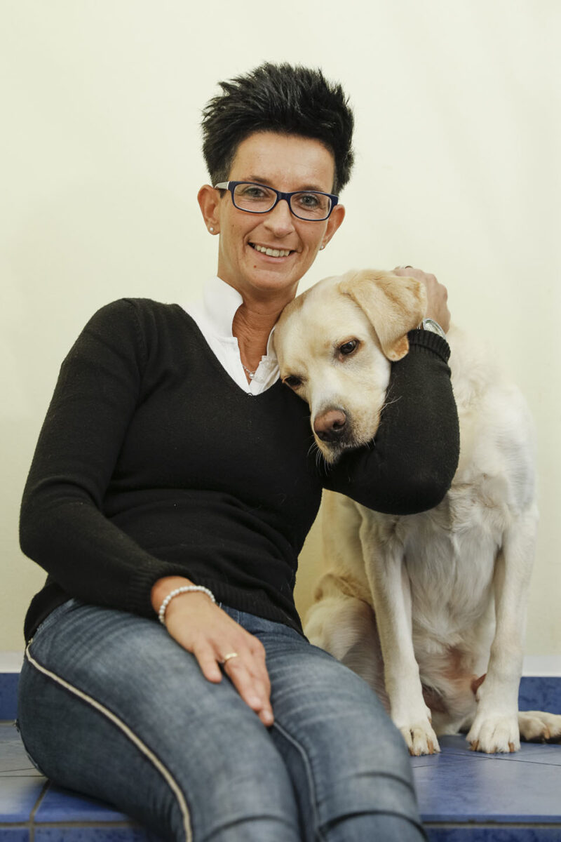 Eine Frau mit einer Brille umarmt einen Hund und blickt in die Kamera.