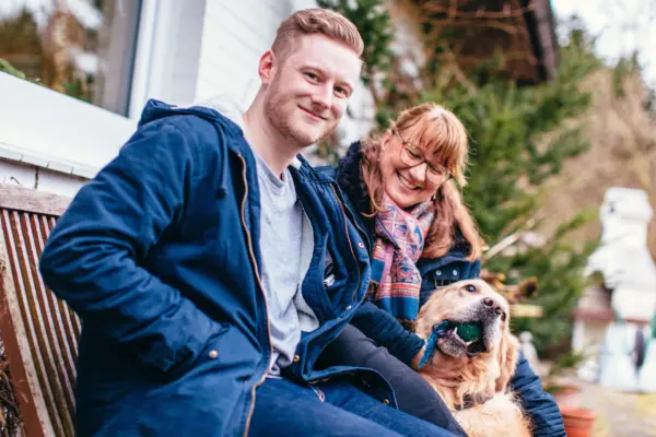 Junger Mann mit einem Bart streichelt einen Hund und sitzt neben einer Frau mit einer Brille.