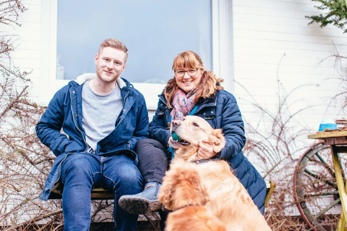 Junger Mann mit einem Bart und eine Frau, die einen Hund streichelt, blicken lächelnd in die Kamera.