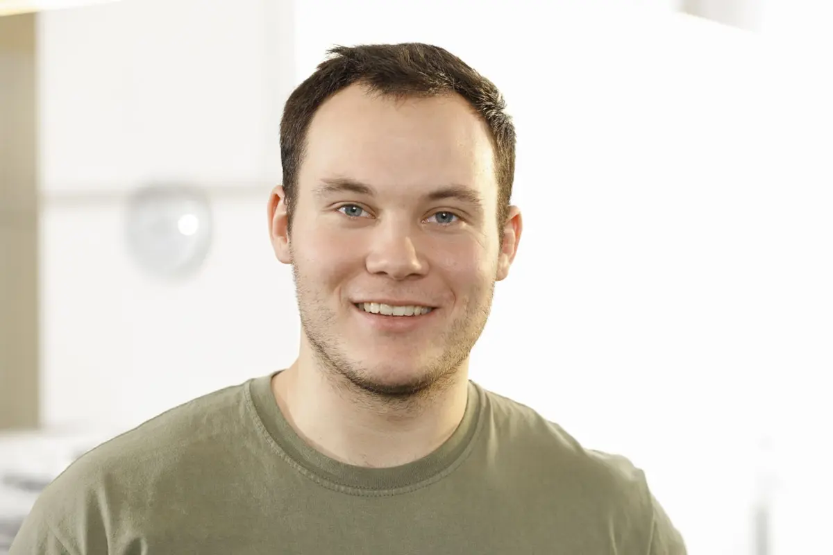 Ein junger Mann mit einem braunen T-Shirt blickt freundlich in die Kamera.