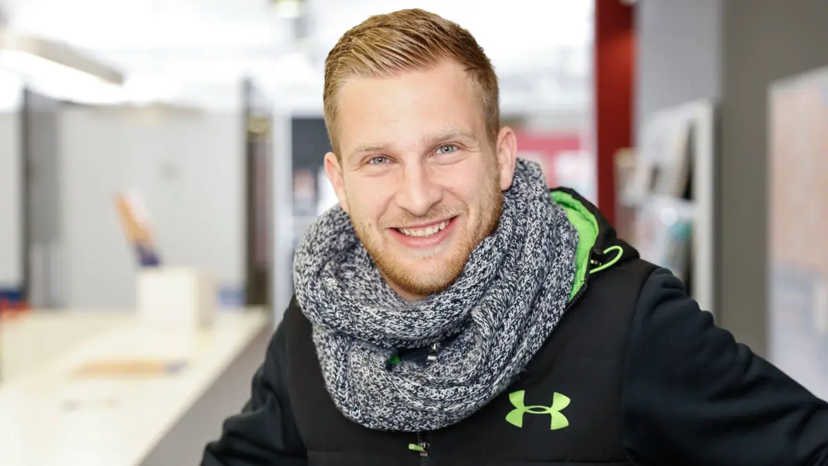Ein junger Mann mit Bart und Schal lächelt in die Kamera.