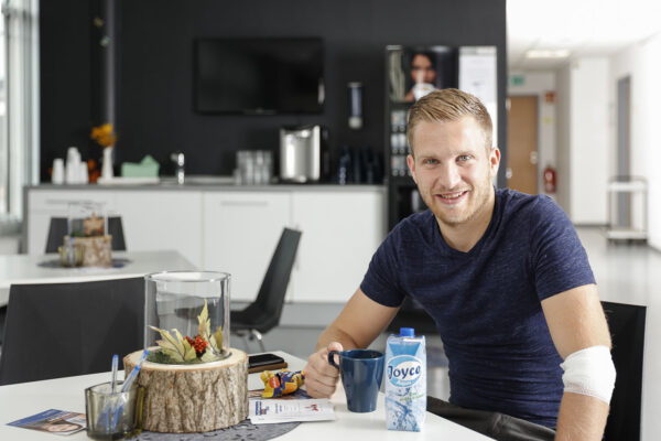 Ein junger Mann mit Bart sitzt an einem Tisch und blickt freundlich in die Kamera.