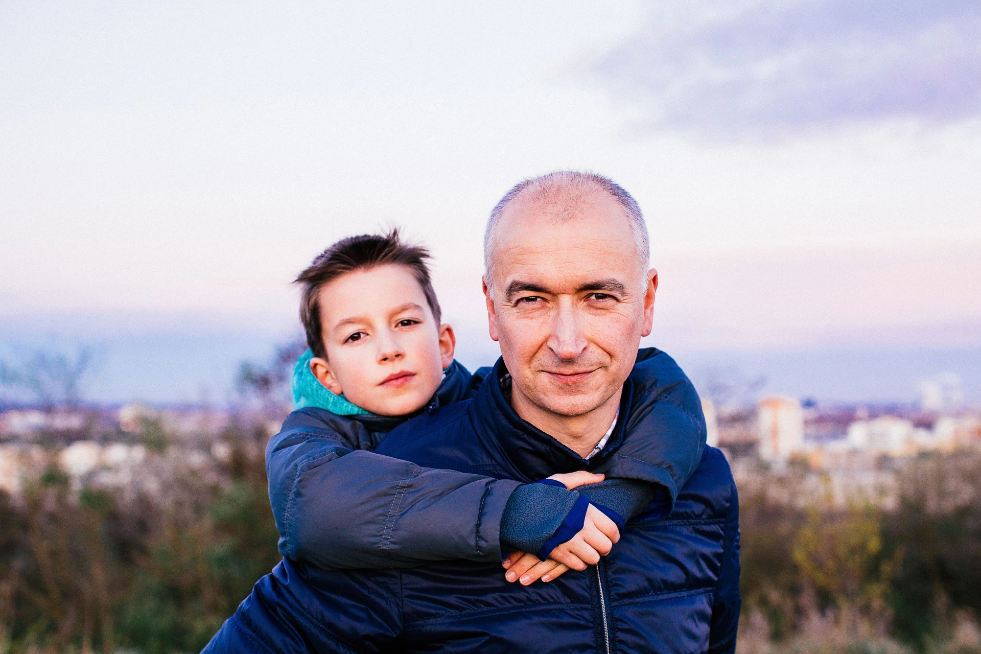 Ein Junge und ein Mann blicken in die Kamera. Dabei hat der Mann sein Sohn auf dem Rücken.